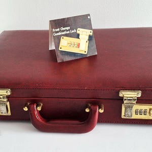 Puede incluir: Un maletín rectangular de color burdeos con asa a juego y herrajes dorados. Cuenta con dos cerraduras de combinación en la parte frontal. Una pequeña tarjeta informativa situada encima muestra una cerradura dorada y el texto Front Change Combination Lock.