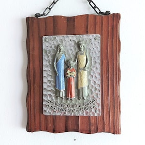 Może przedstawiać: Drewniana tabliczka z metalowym wkładką przedstawiającą rodzinę złożoną z trzech osób. Rodzina jest wykonana z metalu i pomalowana na niebiesko, czerwono i żółto. Tabliczka ma łańcuch do zawieszenia i tekst "Unis dans le même amour nous partageons leur vie".