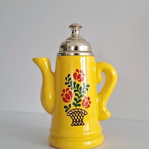 Puede incluir: Una tetera de cerámica de color amarillo brillante con acabado satinado, asa curva y pico corto. La parte frontal está decorada con un diseño pintado de una cesta marrón llena de hojas verdes y tres flores rojas. Tiene una tapa de color plata.