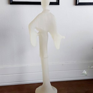 Op de afbeelding: Een wit keramisch beeld van een persoon in een vloeiende jurk. Het beeld staat op een voetstuk en heeft een minimalistisch ontwerp.