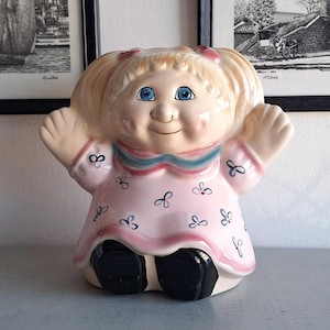 Peut inclure: Une figurine de poupée en céramique avec une robe rose et des yeux bleus. La poupée a des tresses blondes et porte des chaussures noires. La poupée est assise sur une surface gris clair.