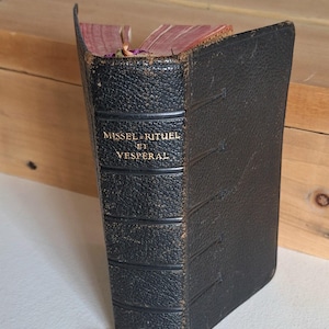 May include: A vintage book with a black leather cover, titled "Missel Rituel et Vesperal" in gold lettering. The book's pages are visible, with a reddish-brown hue. The book is positioned on a white surface, with a wooden box in the background.
