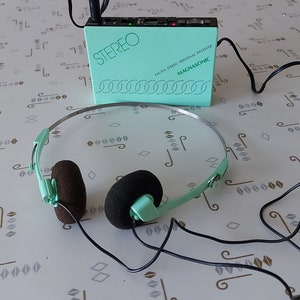 May include: A teal and black AM/FM stereo personal receiver with the text "STEREO" and "MAGNASONIC" on the front. The receiver is connected to a pair of black headphones with teal earcups.