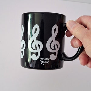 Peut inclure: Une tasse en céramique noire avec des motifs de clés de sol blanches et le texte "Mostly Music" au fond.
