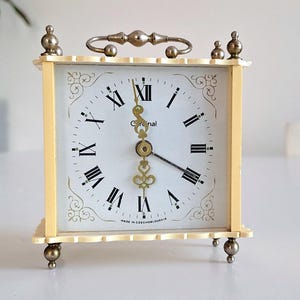 May include: A vintage square clock with a cream-colored frame and a white face. The clock features black Roman numerals, decorative gold accents, and a handle on top. The clock is approximately 5 inches tall.
