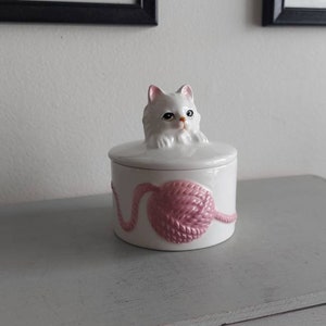Peut inclure: Boîte à bijoux en céramique blanche avec une figurine de chat blanc sur le couvercle. Le chat regarde vers la droite et a du fil rose sur la boîte.