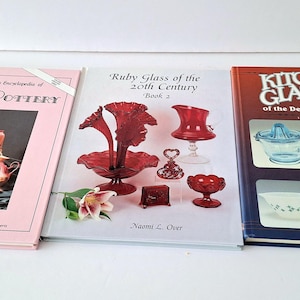 Może przedstawiać: Trzy książki o zabytkowych szkłach i ceramice. Pierwsza książka jest różowa i zawiera zdjęcie Hull Pottery. Druga książka dotyczy rubinowego szkła z XX wieku i zawiera zdjęcie czerwonej szklanej wazony. Trzecia książka jest niebieska i zawiera zdjęcie naczyń kuchennych z okresu Wielkiego Kryzysu.