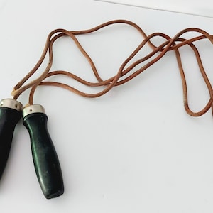Peut inclure: Une corde à sauter en cuir vintage avec des poignées en bois. La corde est brune et les poignées sont noires avec des accents argentés.