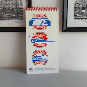 May include: A white notepad with a red spiral binding features three different Molson Export logos, each with a different car design. The logos are blue, red, and gold. The notepad also includes the company's address and phone number.