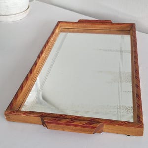 May include: A rectangular serving tray with a mirrored base and a wooden frame. The frame features a carved, rope-like pattern with red painted accents. The tray includes two integrated wooden handles on the shorter sides and a reflective surface.