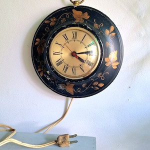 May include: A black and gold vintage electric clock with a floral design. The clock has a round face with Roman numerals and a gold cord.