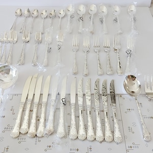May include: A set of silver cutlery, including forks, knives, spoons, and serving pieces, in individual plastic packaging. The cutlery has a decorative, textured design.