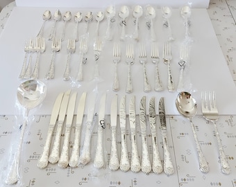 Könnte beinhalten: Ein Set aus silbernem Besteck, bestehend aus Gabeln, Messern, Löffeln und Servierstücken, in einzelnen Plastikverpackungen. Das Besteck hat ein dekoratives, strukturiertes Design.