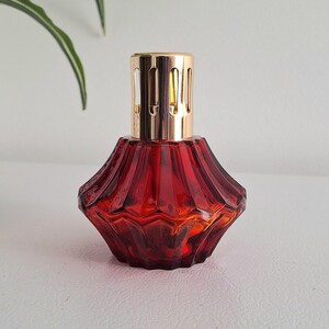 Puede incluir: Lámpara de fragancia decorativa de vidrio rojo con una parte superior de metal dorado. La lámpara tiene un diseño geométrico facetado. La lámpara se utiliza probablemente para aromaterapia o fragancia para el hogar.