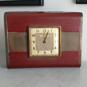 Puede incluir: Un reloj de viaje rojo vintage con esfera cuadrada. El reloj tiene un marco dorado y una esfera beige con números negros. Una banda horizontal de metal cepillado acentúa el exterior rojo similar al cuero.