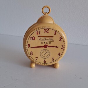 May include: A vintage, cream-colored alarm clock with a round face and a small handle on top. The clock face features black numerals and the words "Reliable BANK" in red. The clock has three small feet.