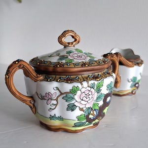 Peut inclure: Un sucrier en céramique blanche avec couvercle et un pot à lait assorti, tous deux ornés de motifs floraux peints à la main en rose, vert et marron. Les pièces présentent des bords festonnés, des anses décoratives et une bordure marron.