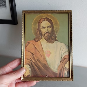 May include: A framed picture of Jesus Christ with a golden halo and a heart with a crown of thorns on his chest. The picture is in a gold frame with a decorative border.
