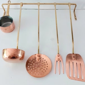 May include: Set of five copper kitchen utensils hanging on a brass rack. The utensils include a ladle, a slotted spoon, a fork, a spatula, and a small saucepan.