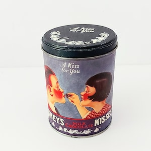 May include: A vintage tin container for Hershey's Kisses milk chocolate. The tin is purple with a black lid and features a retro illustration of two children sharing a kiss with the text "A Kiss for You".