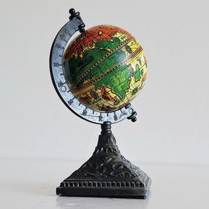 Puede incluir: Un pequeño globo terráqueo con un mapa del mundo colorido, montado sobre un soporte de metal negro. El globo presenta tonos de verde, amarillo, naranja y rojo. El soporte tiene un diseño decorativo y ornamentado.