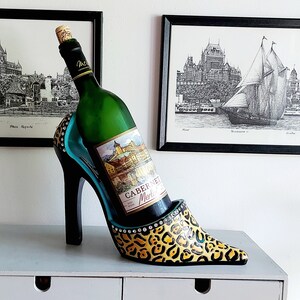 Peut inclure: Un porte-bouteille en forme de chaussure à talon haut noir et turquoise avec un motif léopard et des strass. Une bouteille de vin blanc est dans le porte-bouteille.