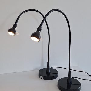 Puede incluir: Dos lámparas de escritorio negras con brazos flexibles de cuello de cisne y bases redondas. Cada lámpara cuenta con un cabezal pequeño y ajustable que emite una luz brillante y enfocada. Las lámparas están conectadas por cables negros y tienen un acabado brillante.