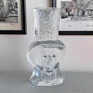 Puede incluir: Un jarrón de vidrio transparente con forma de persona que lleva un sombrero. El jarrón tiene una superficie texturizada y una cara con ojos, nariz y boca.