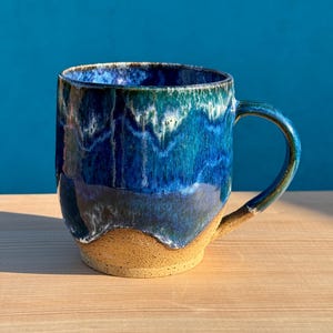 May include: A ceramic mug featuring a glossy glaze with deep blue, teal, and white hues that drip down toward the base. The bottom portion of the mug is made of unglazed, speckled tan clay, providing a textured contrast to the smooth, colorful upper section.