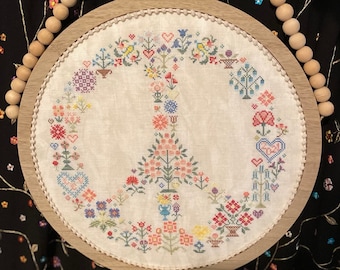 May include: A white embroidery hoop with a peace sign design featuring colorful flowers and hearts. The hoop is made of wood and has a natural finish.