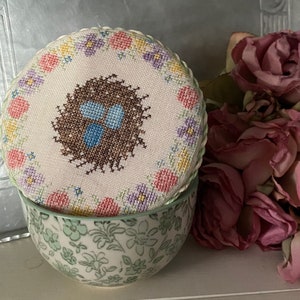 Könnte beinhalten: Eine mit weißem Stoff bezogene Schachtel mit floralem Rand und einem braunen Nest mit blauen Eiern in der Mitte. Die Schachtel steht auf einer grün-weiß gemusterten Schale.