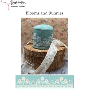 Puede incluir: Un cojín de alfileres azul verdoso con bordados blancos de flores y casas. El cojín de alfileres está sentado en una cesta de mimbre con ribete de encaje blanco. La imagen también incluye un primer plano del patrón de bordado, que presenta flores, casas y un patrón repetitivo. El texto "Blooms and Bunnies" y "No. 00028" también son visibles en la imagen.