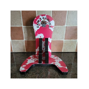 May include: A pink and white gaming controller stand with a black base and two black slots for controllers. The stand has a logo that says "Cam's Gaming" with a skull and crossbones design.