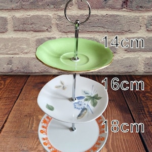 May include: Three-tiered cake stand with a green top tier, a white middle tier with a floral design, and a white bottom tier with an orange floral design. The stand is 14 cm in diameter at the top, 16 cm in diameter in the middle, and 18 cm in diameter at the bottom.