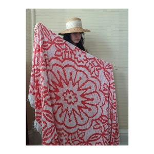 May include: A red and white throw blanket with a large floral pattern. The blanket has a fringe border and is being held up by a person wearing a straw hat.