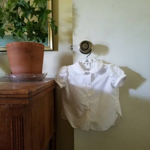May include: A white, short-sleeved blouse with a rounded collar and button-down front hangs on a hanger. The blouse is made of a lightweight fabric. The blouse is hanging on a door.