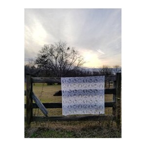 May include: A white crocheted blanket with a blue pattern of circles hanging on a wooden fence against a cloudy sky.