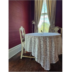 May include: A white lace tablecloth covers a round table with two white chairs. The table is set with a single vase of flowers. The room has dark red walls and a window with white curtains.