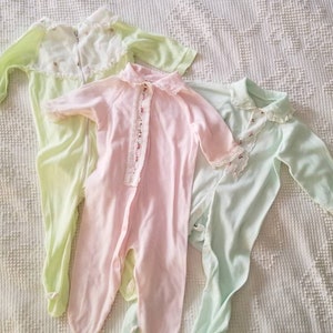 May include: Three vintage baby sleepers in pastel colors: light green, pink, and mint green. Each sleeper has long sleeves, a collar, and a front closure. The pink sleeper has a floral lace detail.