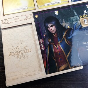 May include: Aeon's End board game component featuring a character card with an illustration of a dark-haired person with blue eyes wearing a red and blue robe. The card has the text "Xaxos, Apprentice Mage" and "2" in a gold circle. There are five red crystals on the card.