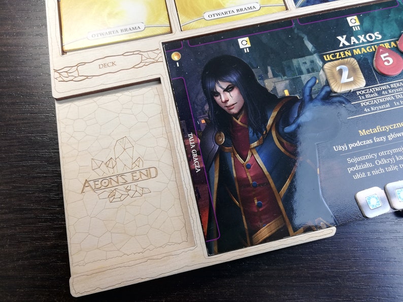 May include: Aeon's End board game component featuring a character card with an illustration of a dark-haired person with blue eyes wearing a red and blue robe. The card has the text "Xaxos, Apprentice Mage" and "2" in a gold circle. There are five red crystals on the card.