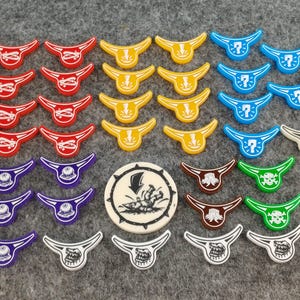 Puede incluir: Una colección de fichas de juego coloridas en rojo, amarillo, azul, morado, marrón, verde y blanco. Las fichas tienen varios diseños, incluyendo cuernos, signos de interrogación y calaveras. Una ficha circular central presenta una ilustración en blanco y negro.