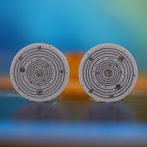 Könnte beinhalten: Zwei weiße, kreisförmige Objekte mit einem schwarzen Sonnensystem-Design. Jede Scheibe zeigt konzentrische Kreise und kleine Planetenillustrationen. Der Hintergrund ist ein verschwommener Farbverlauf aus Blau, Orange und Gelb.