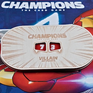 May include: Wooden Champions card game hit point tracker. The tracker displays the numbers 7 and 3 in red boxes. The text "CHAMPIONS THE CARD GAME" and "VILLAIN HIT POINTS" are engraved on the wooden surface.