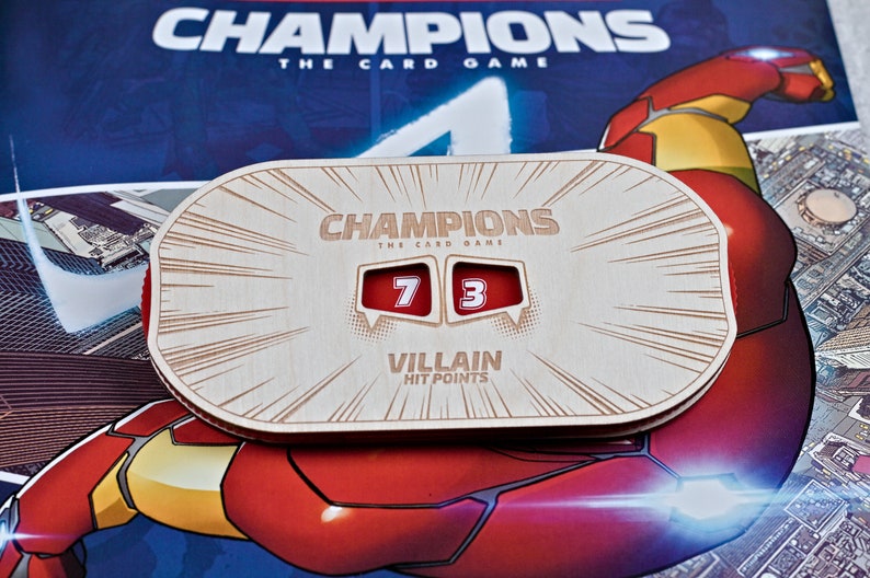 May include: Wooden Champions card game hit point tracker. The tracker displays the numbers 7 and 3 in red boxes. The text "CHAMPIONS THE CARD GAME" and "VILLAIN HIT POINTS" are engraved on the wooden surface.
