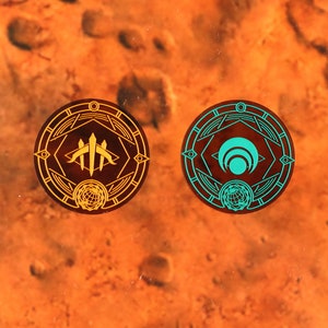 May include: Two circular, decorative, gold and teal colored medallions with intricate designs. The left medallion has a gold background with a teal design of three stylized swords. The right medallion has a teal background with a gold design of a stylized crescent moon.