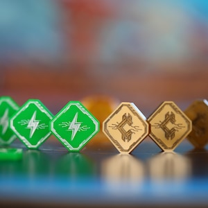 Puede incluir: Primer plano de piezas de juego. Fichas verdes en forma de diamante con un diseño de rayo blanco en primer plano. Fichas de color canela en forma de diamante con un símbolo detrás. El fondo está borroso.