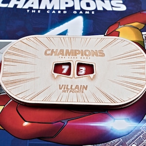 May include: Wooden game accessory for "Champions: The Card Game" with the text "Champions The Card Game" and "Villain Hit Points". The accessory displays the numbers 7 and 3 in red boxes. The background features a comic book illustration.