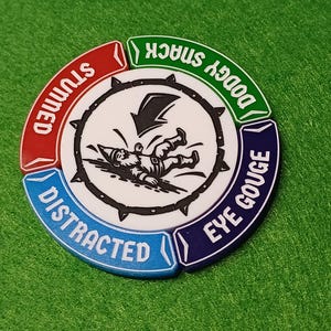 May include: A circular game token with a white center featuring a fallen gnome and a lightning bolt. The token is divided into four sections: red "STUNNED", green "DODGY SNACK", blue "DISTRACTED", and navy "EYE GOUGE".