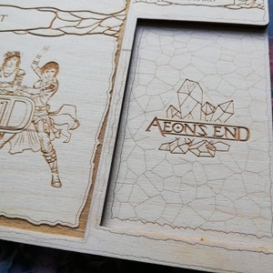 May include: A wooden game board with a light brown finish. The board has a raised section with the words "AEONS END" and a design of crystals. The board is part of a larger game set.